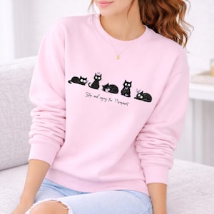 May include: A light pink sweatshirt featuring a row of black cat illustrations with white accents. The text "Stop and enjoy the Moment" is printed below the cats. The sweatshirt has long sleeves and a crew neck.