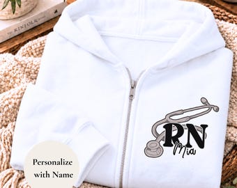 May include: A white zip-up hoodie featuring a graphic of a grey stethoscope and the black text RN Mia. A circular white label on the lower left side contains the black text Personalize with Name. The garment has a hood and a metal zipper.