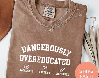 May include: A brown t-shirt with the text "DANGEROUSLY OVEREDUCATED" in white. Below the text are three checkboxes with the text "BACHELOR'S", "MASTER'S", and "DOCTORATE". The shirt is on a light brown surface. The text "COLOR: ESPRESSO" is at the bottom of the image.