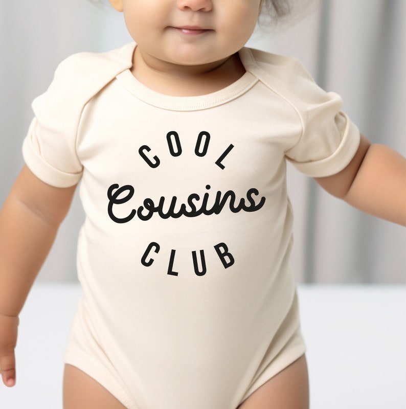 May include: A cream-colored baby onesie with short sleeves and black text that reads "COOL Cousins CLUB".