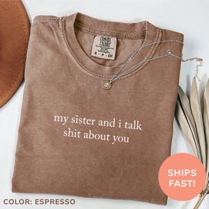 Puede incluir: Camiseta Comfort Colors color espresso con el texto blanco "my sister and i talk shit about you". Un collar plateado con un pequeño colgante descansa sobre la camiseta. La camiseta tiene cuello redondo y una etiqueta pequeña.