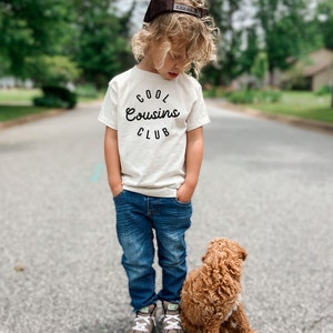 May include: A young boy wearing a white t-shirt with the text "COOL Cousins CLUB" in black lettering, blue jeans, and a brown baseball cap. He is standing next to a small brown dog.