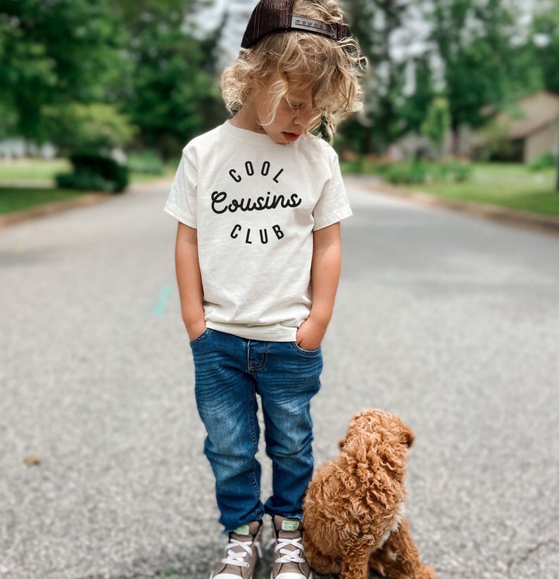 May include: A young boy wearing a white t-shirt with the text "COOL Cousins CLUB" in black lettering, blue jeans, and a brown baseball cap. He is standing next to a small brown dog.