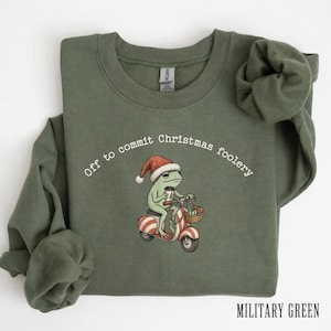 May include: An olive green crewneck sweatshirt featuring a graphic of a frog wearing a Santa hat, riding a red and white striped scooter while holding a coffee cup. The text above the frog reads, Off to commit Christmas foolery.