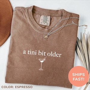 May include: Espresso brown t-shirt with the text "a tini bit older" and a martini glass graphic.