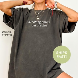 May include: An oversized t-shirt in a dark grey colour with the text "surviving purely out of spite" printed in white on the front. The shirt is made from a soft, comfortable material and has a relaxed fit.