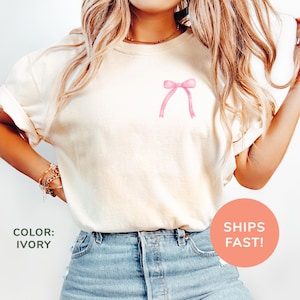 Könnte beinhalten: Ein weißes T-Shirt mit einer rosa Schleife auf der Vorderseite. Das Shirt ist aus einem weichen, bequemen Material und perfekt für den Alltag.