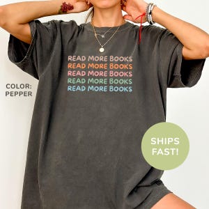 Puede incluir: Camiseta gris oscuro con el texto "READ MORE BOOKS" en colores arcoíris. La camiseta tiene un corte holgado y mangas cortas. El texto "COLOR: PEPPER" está en el lado izquierdo de la camiseta.