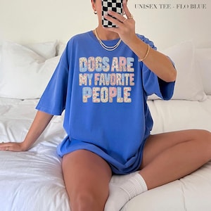 May include: A periwinkle blue short-sleeve t-shirt featuring the text "DOGS ARE MY FAVORITE PEOPLE" in a blocky, multi-colored floral and checkered pattern. The shirt is labeled as "UNISEX TEE - FLO BLUE" in the upper right corner.