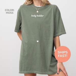 Peut inclure: T-shirt vert mousse surdimensionné avec le texte "body builder" en blanc. Un petit cœur blanc est en dessous du texte. Le t-shirt a des manches courtes et un col rond. Le t-shirt est de couleur unie.