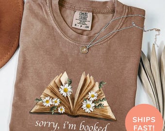 May include: A brown t-shirt with a vintage-style open book design and the text "sorry, i'm booked". The book is decorated with white daisies and green leaves. The shirt is made of a soft, comfortable material and has a distressed look.