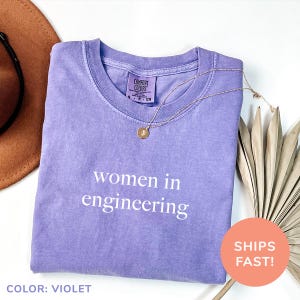 May include: A purple t-shirt with the text "women in engineering" printed in white. The shirt is made of a soft, comfortable fabric.