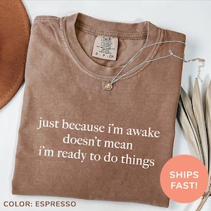 May include: Espresso brown t-shirt with the text "just because i'm awake doesn't mean i'm ready to do things" printed in white. The shirt is made of a soft, comfortable material.