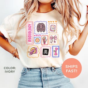 Op de afbeelding: Ivoor kleurig t-shirt met een reeks kleurrijke illustraties in postzegelstijl gerelateerd aan radiologie. De postzegels bevatten afbeeldingen van een ribbenkast, röntgenfoto's en een CT-scanner. Het woord "RADIOLOGY" is verticaal in roze gedrukt.