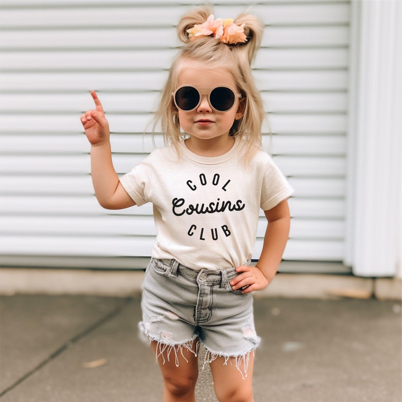 May include: A young child wearing a cream colored t-shirt with the text "Cool Cousins Club" in black. The child is wearing denim shorts and round sunglasses.