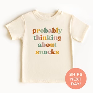May include: Cream-colored short-sleeved t-shirt with the phrase "probably thinking about snacks" in colorful, retro-style lettering. The shirt has a round neckline and a relaxed fit, suitable for children. A peach-colored circle with the text "SHIPS NEXT DAY!" is in the bottom right corner.