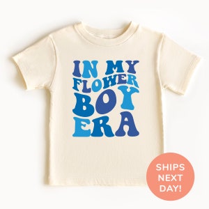 May include: A cream colored t-shirt with blue text that reads "In My Flower Boy Era".