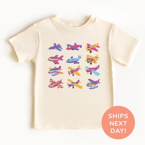May include: Cream-colored t-shirt featuring a colorful array of cartoon airplanes. The planes are in various colors, including purple, pink, yellow, and blue. The shirt has short sleeves and a round neckline. The text "SHIPS NEXT DAY!" is on a round sticker.