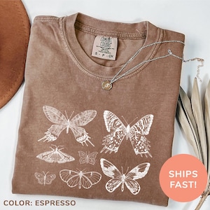 以下が含まれることがあります： ヴィンテージスタイルの蝶の模様が施された茶色のTシャツ。シャツは使い古したような風合いがあり、柔らかく快適な生地で作られています。タグには「COMFORT COLORS」の文字が見えます。シャツは「ESPRESSO」の色で販売されています。