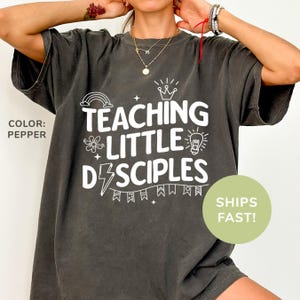 Könnte beinhalten: Pfefferfarbenes T-Shirt mit dem weißen Text "TEACHING LITTLE DISCIPLES". Das Design umfasst einen Regenbogen, eine Blume, eine Krone, eine Glühbirne und einen Blitz. Das Shirt hat einen Vintage-Look.