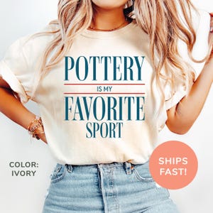 May include: Ivory-colored t-shirt with the text "POTTERY IS MY FAVORITE SPORT" in navy blue. The text is centered and has a red line above and below the words "IS MY". The shirt is paired with blue denim jeans.