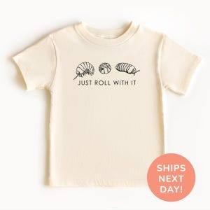 May include: Cream-colored t-shirt featuring three black line-drawn illustrations of pill bugs above the phrase "JUST ROLL WITH IT." The shirt is a short-sleeved crew neck.