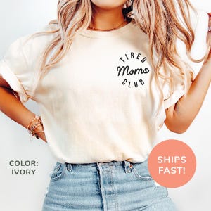 May include: Ivory-colored t-shirt with rolled-up sleeves and the text "Tired Moms Club" in a black circular design. The shirt is paired with blue denim shorts. The color of the shirt is ivory.