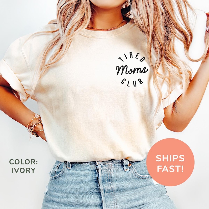 May include: Ivory-colored t-shirt with rolled-up sleeves and the text "Tired Moms Club" in a black circular design. The shirt is paired with blue denim shorts. The color of the shirt is ivory.