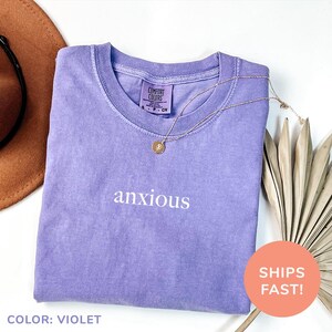 a purple shirt that says anxious next to a hat