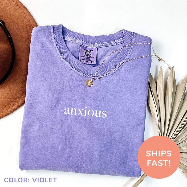 a purple shirt that says anxious next to a hat