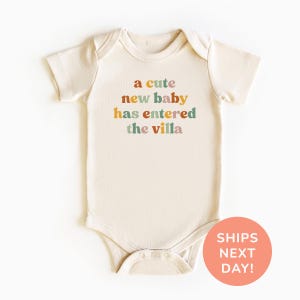 Puede incluir: Body de bebé color crema con el texto "a cute new baby has entered the villa" en letras coloridas. El body tiene mangas cortas y corchetes en la parte inferior. Un círculo color melocotón con el texto "SHIPS NEXT DAY!" está en la esquina inferior derecha.