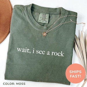 Könnte beinhalten: Ein moosgrünes T-Shirt mit dem Aufdruck "wait, i see a rock" in Weiß. Das Shirt ist aus einem weichen, bequemen Material.