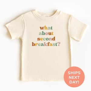 May include: A cream colored short-sleeved t-shirt with the text "what about second breakfast?" printed in colorful letters.