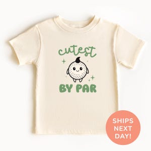 A cream-colored t-shirt with a cartoon golf ball and the text "cutest BY par" printed on it.