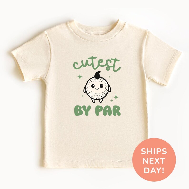 A cream-colored t-shirt with a cartoon golf ball and the text "cutest BY par" printed on it.