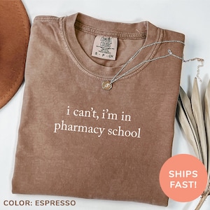 May include: Espresso-colored t-shirt with the text "i can't, i'm in pharmacy school" in white. The shirt has a crew neck and short sleeves. A silver necklace with a small pendant is draped across the shirt.