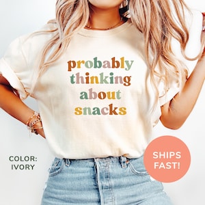 Pode incluir: Camiseta cor marfim com o texto "probably thinking about snacks" em uma fonte retrô. O texto está em tons de marrom, rosa, verde e amarelo. A camiseta é combinada com jeans azuis.