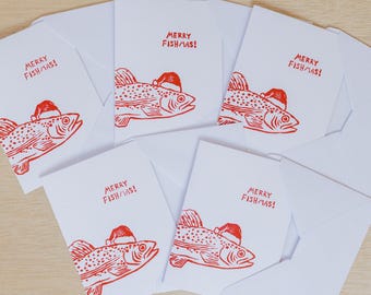 May include: A set of white greeting cards with red illustrations of fish wearing Santa hats. The cards read "MERRY FISH/MAS!" in red text. The cards are accompanied by white envelopes, perfect for holiday greetings.