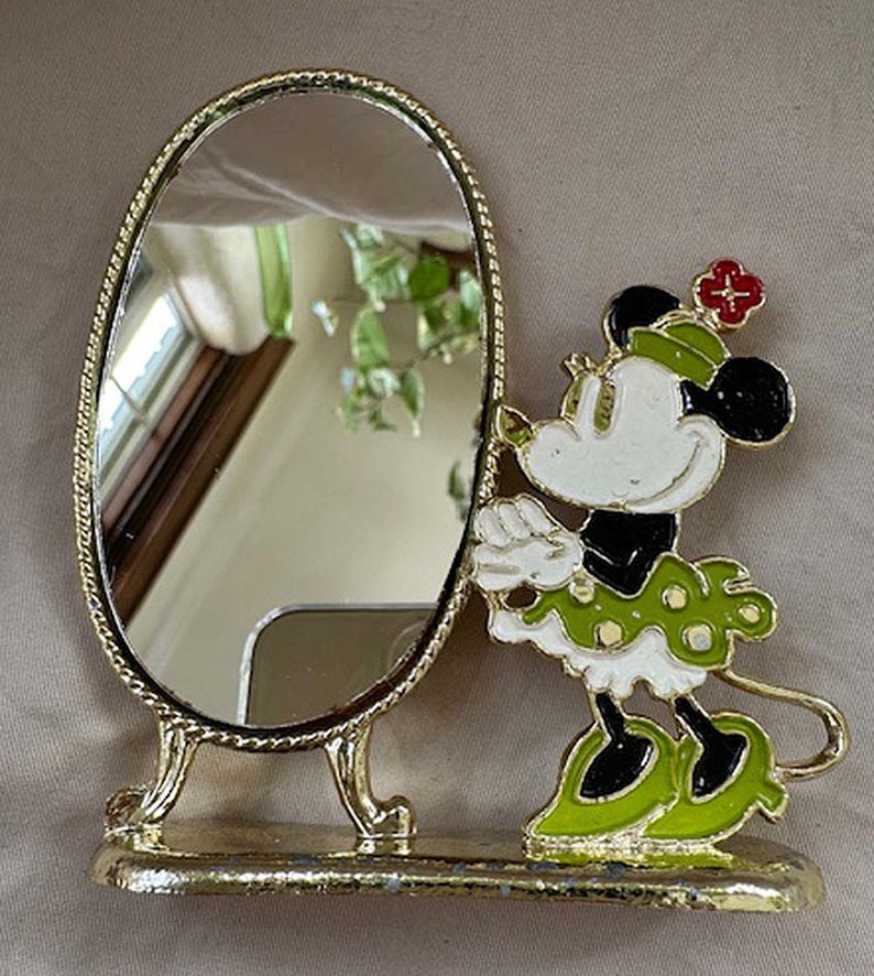 Może przedstawiać: Owalne lusterko kosmetyczne ze złotą ramą i podstawą. Figurka Myszki Minnie w zielono-białej sukience w grochy stoi obok lustra. Figurka ma czerwony kwiat na kapeluszu.