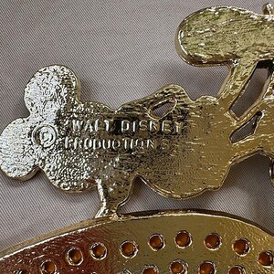 Może przedstawiać: Złoty metalowy element dekoracyjny z wytłoczonym napisem "WALT DISNEY PRODUCTIONS". Element ma kształt głowy Myszki Miki i inne elementy dekoracyjne. Element ma błyszczącą, odblaskową powierzchnię.