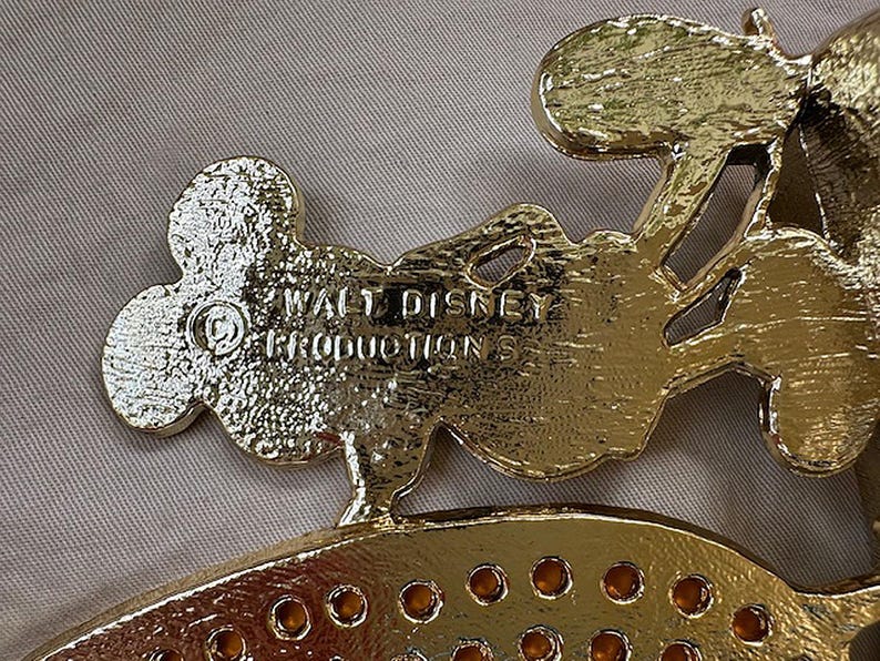 Może przedstawiać: Złoty metalowy element dekoracyjny z wytłoczonym napisem "WALT DISNEY PRODUCTIONS". Element ma kształt głowy Myszki Miki i inne elementy dekoracyjne. Element ma błyszczącą, odblaskową powierzchnię.