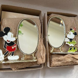 Może przedstawiać: Dwa zabytkowe lustra dekoracyjne z Myszka Miki i Minnie. Każde lustro ma złotą ramę i podstawę. Miki ma na sobie czerwone szorty, a Minnie zieloną sp&oacute;dnicę. Lustra są w oryginalnych pudełkach.