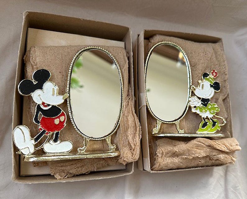 Może przedstawiać: Dwa zabytkowe lustra dekoracyjne z Myszka Miki i Minnie. Każde lustro ma złotą ramę i podstawę. Miki ma na sobie czerwone szorty, a Minnie zieloną sp&oacute;dnicę. Lustra są w oryginalnych pudełkach.