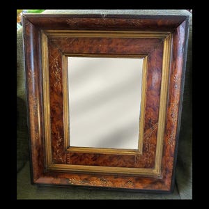 May include: Rectangular mirror with a dark brown, patterned wooden frame. The frame has a gold inner border and decorative gold leaf accents. The mirror's surface is reflective, and the overall design is classic and elegant.