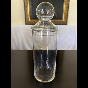 May include: A clear glass apothecary jar with a cylindrical body and a matching glass lid featuring a prominent spherical knob handle. The container is transparent, smooth, and features a simple, functional design with a circular rim and base.
