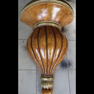 May include: Ornate, wall-mounted shelf in shades of brown and gold. The shelf has a textured, rounded top and a bulbous, ribbed body. Gold trim accents the edges and base, creating a decorative, vintage aesthetic.