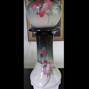 May include: A white ceramic planter with pink and green floral designs sits on a black ceramic pedestal. The planter has a rounded shape and a scalloped base.