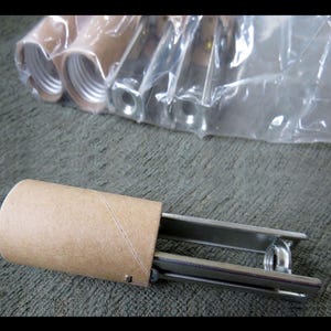 May include: A close-up of a cardboard tube with a metal cap on one end. The tube is partially inserted into a metal device with a silver finish. The device has a small metal ring on the end.