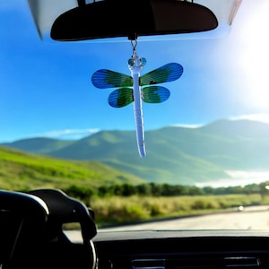 May include: A blue and green dragonfly car charm hanging from the rearview mirror of a car. The dragonfly is made of glass and has a white body. The car is driving on a road with green hills in the background.