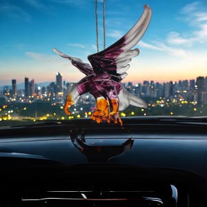 Può includere: Un ornamento per auto in vetro viola e trasparente a forma di aquila appeso a uno specchietto retrovisore. L'aquila è in volo con le ali spiegate. Lo sfondo è uno skyline urbano al tramonto.